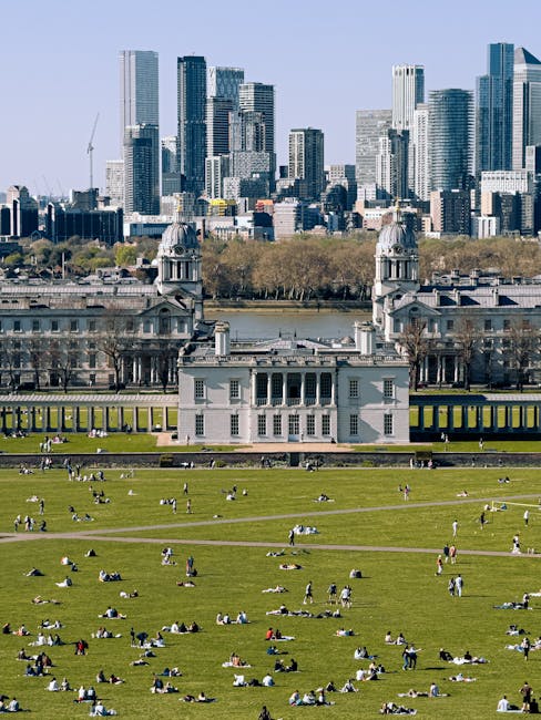 A large public park with expansive green lawns is occupied by numerous people sitting or lying on the grass, many of whom are socialising or relaxing in groups or individually. The grass appears well-maintained and vibrant, with some areas showing pathways or walking routes. In the background, there is a stately, historic building with classical architectural features, including columns, decorative stonework, and two prominent towers topped with domes. Behind this building, a river runs horizontally across the scene, with trees lining its banks. Beyond the river, a modern city skyline with a variety of tall, glass-clad skyscrapers dominates the horizon, reflecting light and creating a contrasting backdrop to the foreground. The overall scene is well-lit with natural daylight, suggesting a clear, calm day. The image exemplifies the urban environment where outdoor leisure activities take place amidst significant historic and modern architectural elements, similar to how private property or outdoor spaces are maintained for recreational use. Waste Clearance Greenwich could facilitate clearing waste generated during outdoor events or gatherings from such public spaces, supporting alternative waste handling outside municipal collection services.
