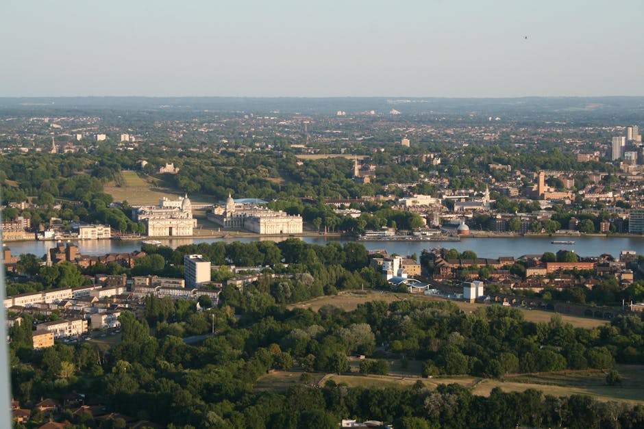 An aerial view of a cityscape showing a wide river flanked by historic and modern buildings, with a prominent castle-like structure on the riverbank surrounded by lush green trees. The foreground features a mix of residential and commercial properties with visible rooftops and roads, while the background extends into a distant horizon with urban sprawl and patches of greenery. The scene is bathed in natural daylight, emphasizing the contrast between the dense greenery and the built environment, highlighting the importance of managing waste and rubbish removal to maintain such urban landscapes. This landscape suggests the significance of alternative waste handling methods, such as private rubbish collection services, in preserving the city's aesthetic and environmental health, aligning with services like those offered by Waste Clearance Greenwich, particularly around areas with historic sites and dense city areas requiring efficient waste clearance solutions.