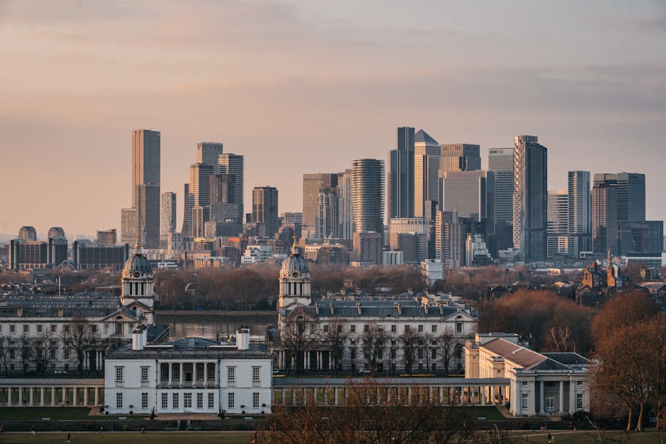 A daytime view of a city skyline with a cluster of modern high-rise office buildings and residential towers in the background, exhibiting glass facades that reflect the daylight. In the foreground, there is a historic white building with classical architectural features, including dome-shaped cupolas topped with decorative finials, arched windows, and a symmetrical layout. The landscape around the historic building includes leafless trees and manicured lawns, suggesting late autumn or early winter season. The sky above the city contains soft, pastel-hued clouds, indicating a calm and clear day. This scene depicts an urban area where private waste clearance services, such as those offered by Waste Clearance Greenwich, might be relevant for managing rubbish removal from historic or commercial properties within a busy metropolitan environment, providing an alternative or supplementary solution to local authority waste collection options.