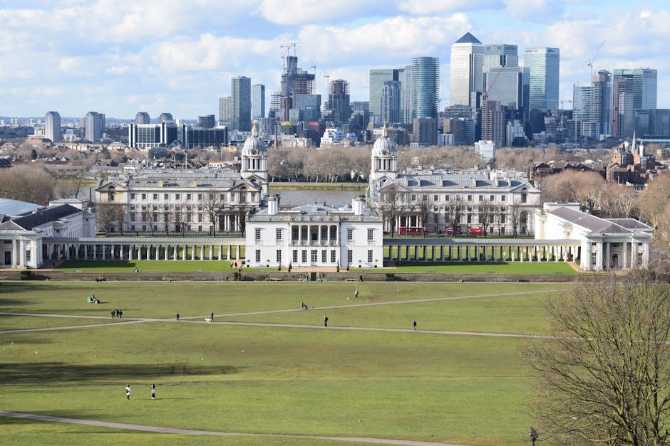 A wide view of a large historic white building set in a landscaped garden with green lawns, pathways, and a terrace area, situated in an urban park environment. The building features symmetrical architecture with multiple windows, ornate towers, and a colonnaded façade, with a long colonnade extending to both sides. In the foreground, the expansive lawn is dotted with small groups of people walking or relaxing. In the background, there is a dense cluster of modern high-rise office buildings and skyscrapers under a partly cloudy sky, contrasting with the classical architectural style of the historic structure. The scene captures a mix of historic and contemporary city elements, with the landscaped park in the foreground providing a tranquil setting amid the urban skyline. The lighting appears natural, suggesting daytime conditions with some cloud cover affecting the overall brightness, and the environment is a well-maintained public space near the city center, relevant to discussions of urban parks, historic landmarks, or local infrastructure.