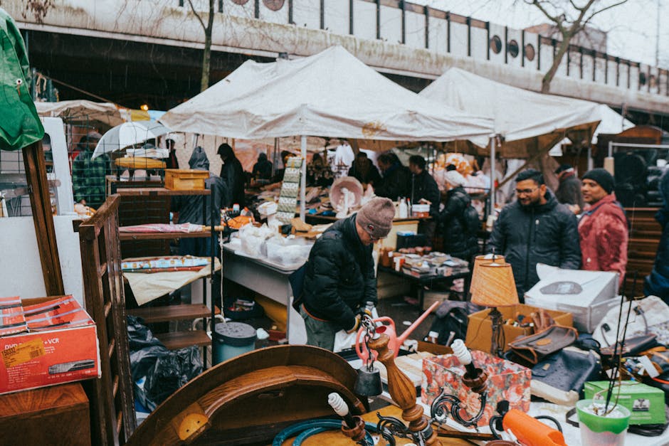 A busy outdoor market scene under overcast weather, featuring multiple white tents with scalloped edges set up along a paved area, possibly on a street or marketplace in Greenwich. The foreground shows various household items and small furniture, including a wooden chair, a table lamp with a yellow shade, and eclectic decorative objects, arranged clutteredly on tables and the pavement. Several individuals are visible, browsing and examining goods; one person in a gray knit hat and dark coat is focused on a piece of furniture or object, while others stand nearby, engaging with vendors. The background reveals more market stalls with additional merchandise, some shaded by umbrellas or additional tents, and a concrete bridge with railing, elevated above the scene. The overall lighting appears diffused due to the cloudy sky, emphasizing the casual, communal atmosphere typical of on-site clearance or private sale environments, where waste clearance services like Waste Clearance Greenwich might be involved in clearing residual rubbish or surplus items after market events.