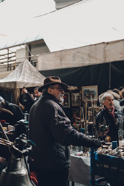 A man wearing a dark jacket and wide-brimmed hat stands at an outdoor market stall, examining various items on display. The scene is set under a large canopy that provides shelter, with other market visitors visible in the background, some browsing and others engaging with vendors. The stall features a collection of objects, possibly antiques or collectibles, arranged on a table covered with a cloth. Surrounding the man are additional market stalls and displays, creating a busy, lively marketplace environment. The lighting suggests natural daylight, with ambient shadows cast underneath the canopy. This marketplace scene illustrates a typical environment where independent collection and on-site clearance of vintage or used goods are common, often involving private vendors handling their own disposal rather than relying on local authority rubbish collection services. Waste Clearance Greenwich and similar companies might be involved in clearing unsold items or waste generated by such markets, supporting alternative waste handling methods in these settings.