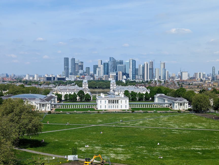 A wide view of Greenwich Park during daylight shows a large, historic white building with symmetrical architectural details, including domed towers and colonnades, situated on a well-maintained green lawn with walking paths crossing the open space. In the foreground, there is a small construction excavator parked on the grass, indicating ongoing work or clearance activities. The park is bordered by a row of mature trees, adding a lush, natural border to the scene. Beyond the park, the modern city skyline of tall skyscrapers and office buildings rises prominently, contrasting with the classical architecture below. The sky is mostly clear with a few scattered clouds, providing bright natural light that highlights both the historic structure and the cityscape. This image subtly illustrates the ongoing maintenance or clearance efforts typical of independent waste management services, such as those provided by Waste Clearance Greenwich, supporting private rubbish removal within historic and urban settings.