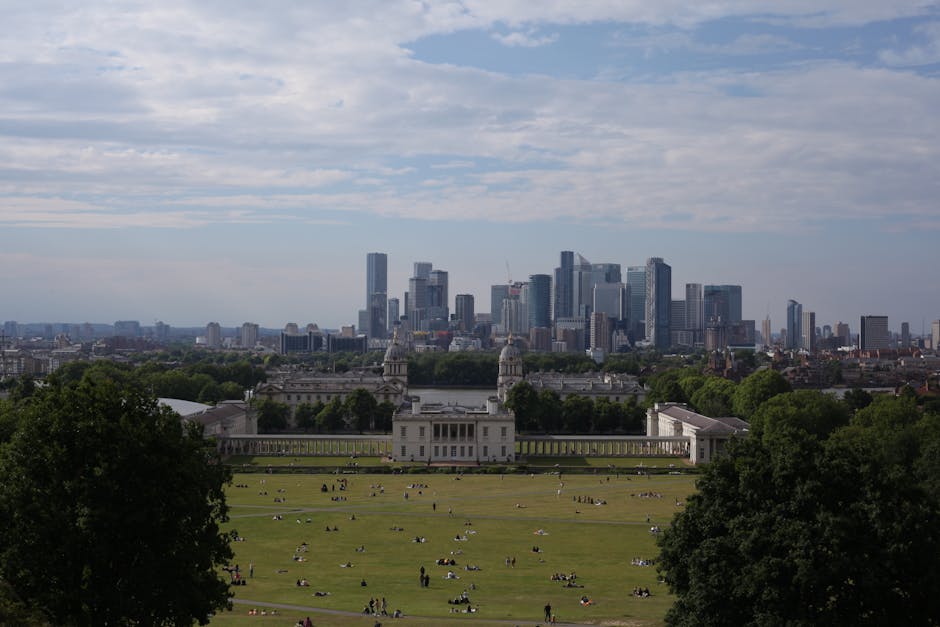 A wide view of Greenwich Park during daylight shows a large, historic white building with symmetrical architectural details, including domed towers and colonnades, situated on a well-maintained green lawn with walking paths crossing the open space. In the foreground, there is a small construction excavator parked on the grass, indicating ongoing work or clearance activities. The park is bordered by a row of mature trees, adding a lush, natural border to the scene. Beyond the park, the modern city skyline of tall skyscrapers and office buildings rises prominently, contrasting with the classical architecture below. The sky is mostly clear with a few scattered clouds, providing bright natural light that highlights both the historic structure and the cityscape. This image subtly illustrates the ongoing maintenance or clearance efforts typical of independent waste management services, such as those provided by Waste Clearance Greenwich, supporting private rubbish removal within historic and urban settings.