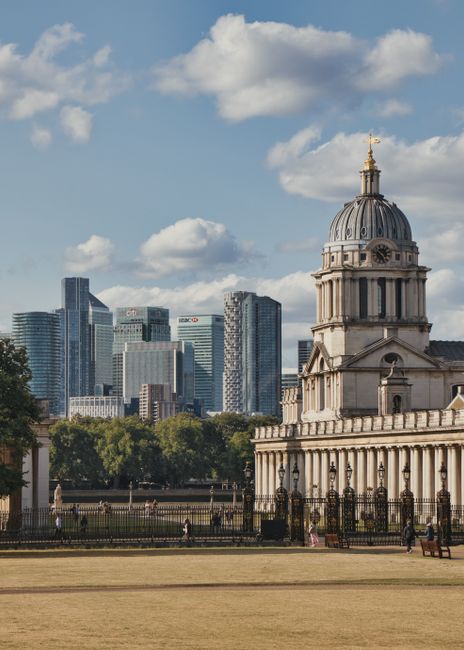 The image shows a historic stone building with a prominent domed tower featuring classical architectural elements, including columns and decorative moldings, situated in the foreground along a paved walkway. The building's light-colored stone façade contrasts with the modern cityscape of high-rise office buildings and skyscrapers, which are visible in the background under a partly cloudy sky. The scene appears to be taken in a public urban park or open space, with a low fence surrounding the historic building and a few park visitors walking or sitting nearby. The green trees and grass area are also partly visible behind the fence, adding natural elements to the setting. The overall atmosphere combines Victorian architecture with contemporary city development, illustrating a typical scene where heritage structures coexist with modern commercial buildings, relevant to discussions of independent or alternative waste handling options within urban environments. The lighting is natural, suggesting daytime with bright but diffuse sunlight highlighting details on both historic and modern structures, supporting the context of property upkeep and preservation often associated with site clearance or rubbish removal services by companies such as Waste Clearance Greenwich.