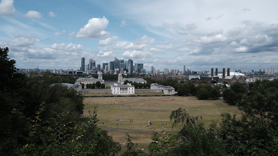 A wide view of a city skyline showing modern high-rise buildings and skyscrapers situated behind a historic white mansion with classical architecture, columns, and a long surrounding wall, set on a large grassy area with scattered people relaxing and walking. The foreground features green trees and bushes framing the scene, while the background sky is partly cloudy with patches of blue visible through the clouds. The image captures an urban landscape that might be associated with private or independent waste management activities, supporting on-site clearance or rubbish removal services in the area. The outdoor setting emphasizes the contrast between the natural environment and the dense city development beyond, demonstrating the kind of surroundings where rubbish clearance could be necessary for maintaining aesthetics in residential or public spaces, consistent with services offered by Waste Clearance Greenwich.