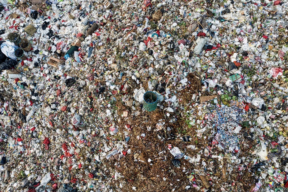 An expansive view of a large landfill site filled with mixed waste materials, including plastic bottles, food packaging, paper, cardboard, and organic refuse. The waste is densely piled, with various colors such as red, blue, white, brown, and green, indicating different types of trash. In the midst of the debris, a small plastic container and scattered pieces of broken wood can be seen, with some areas showing signs of soil and organic matter. The pile extends across the entire image, with no clear pathways visible, and the waste appears to be in a natural outdoor environment, possibly a designated disposal area. The lighting is natural, with a diffuse light source suggesting an overcast day, emphasizing the chaotic and unorganized nature of the waste. This scene underscores the importance of proper rubbish management and disposal services, such as those offered by Waste Clearance Greenwich, which specializes in independent waste clearance and private disposal options for residents and businesses in the SE10 area of Greenwich.